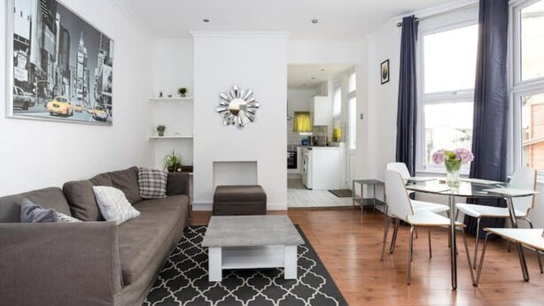 Un salon moderne avec un canapé gris, une table basse et un coin repas comprenant une table et des chaises en verre. On aperçoit une cuisine au fond, une grande fenêtre avec des rideaux et un tableau représentant un paysage urbain accroché au mur.
