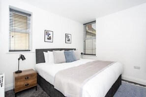 A bedroom with a bed, a nightstand, a lamp, and two windows with blinds.