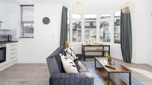 Een moderne woonkamer met een bank, een salontafel en een eettafel. Er is een keukenhoek met een magnetron en een raam met gordijnen.