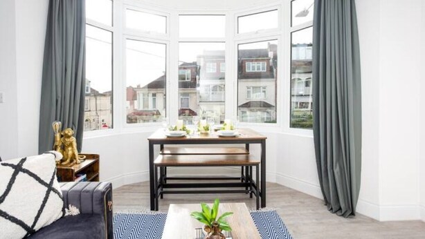 Een hoekkamer met erker, een eettafel gedekt met borden en glazen, een stoel met een gestreept kussen en een klein beeldje op een bijzettafeltje.