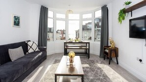 A modern living room with a black sofa, a wooden coffee table, a TV mounted on the wall, and a large window offering a view of the city. - Trendy 2 Bed Flat, close to Beach & Restaurants (Southend-on-Sea)