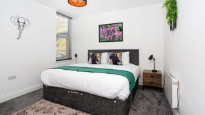 A bedroom with a bed, a bedside table, a lamp, a radiator, a window with blinds, a wall art of an elephant, a neon sign, and a hanging plant. - Trendy 2 Bed Flat, close to Beach & Restaurants (Southend-on-Sea)