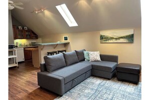 A modern living room with a sectional sofa, a small ottoman, a painting on the wall, and a skylight above the kitchen area. - Private, great loc - ideal for long / short stay! (Canton)