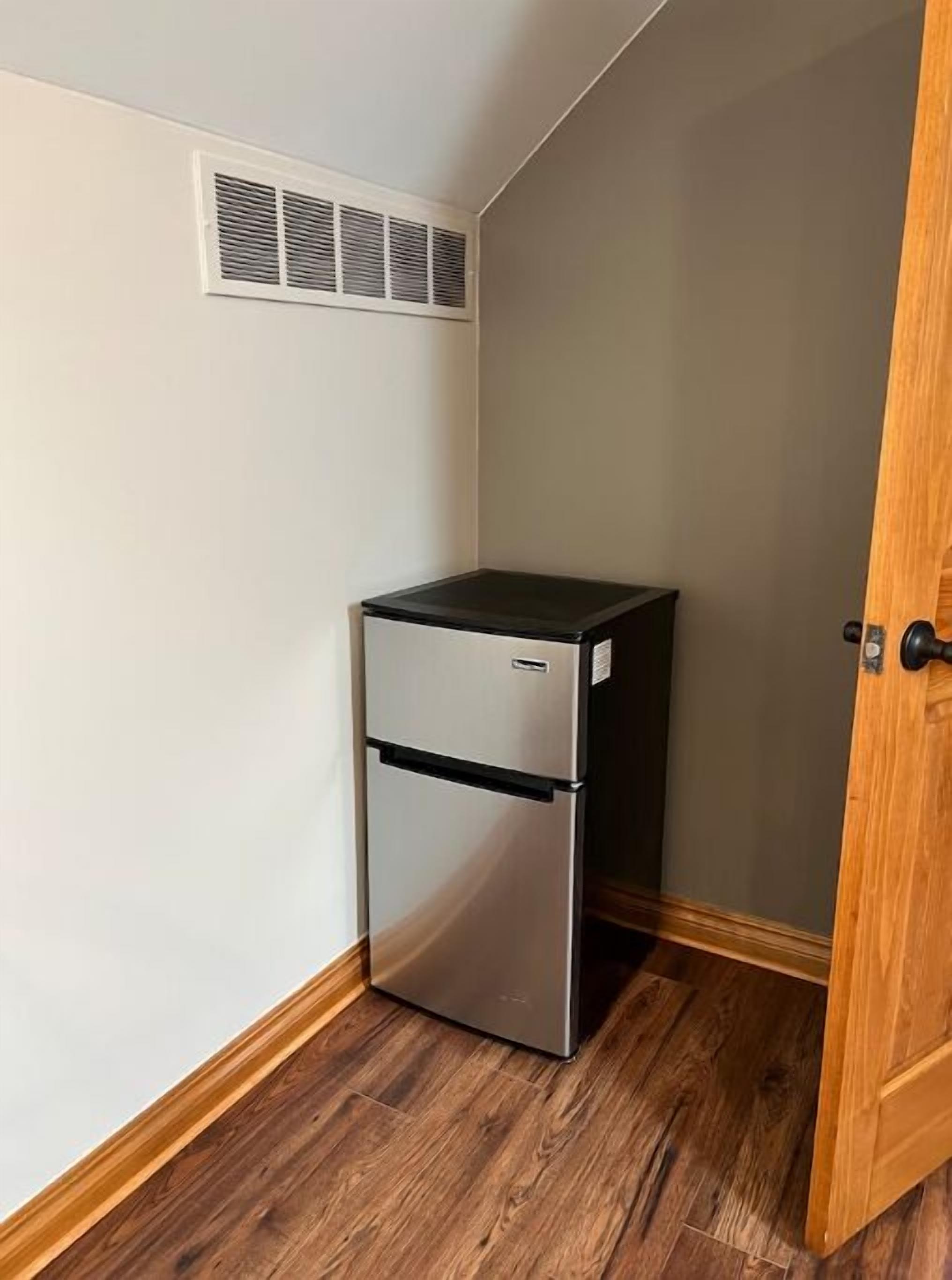 A compact refrigerator in a corner with a wooden floor and a door to the right.