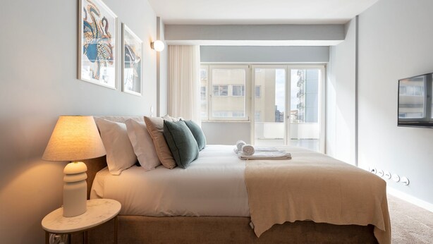 Ein Hotelzimmer mit Bett, einer Nachttischlampe, einem Fenster mit Blick auf Gebäude und Kunstwerken an der Wand.
