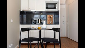 A compact kitchen with a microwave, coffee maker, and a wooden clock on the wall. There is a small table with two chairs and a vase.