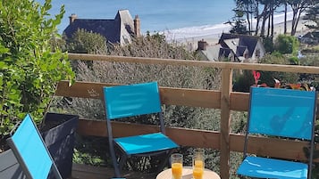 Une terrasse en bord de mer, avec des chaises bleues et une table où sont disposées des boissons, offrant une vue sur l’océan et une ville côtière.