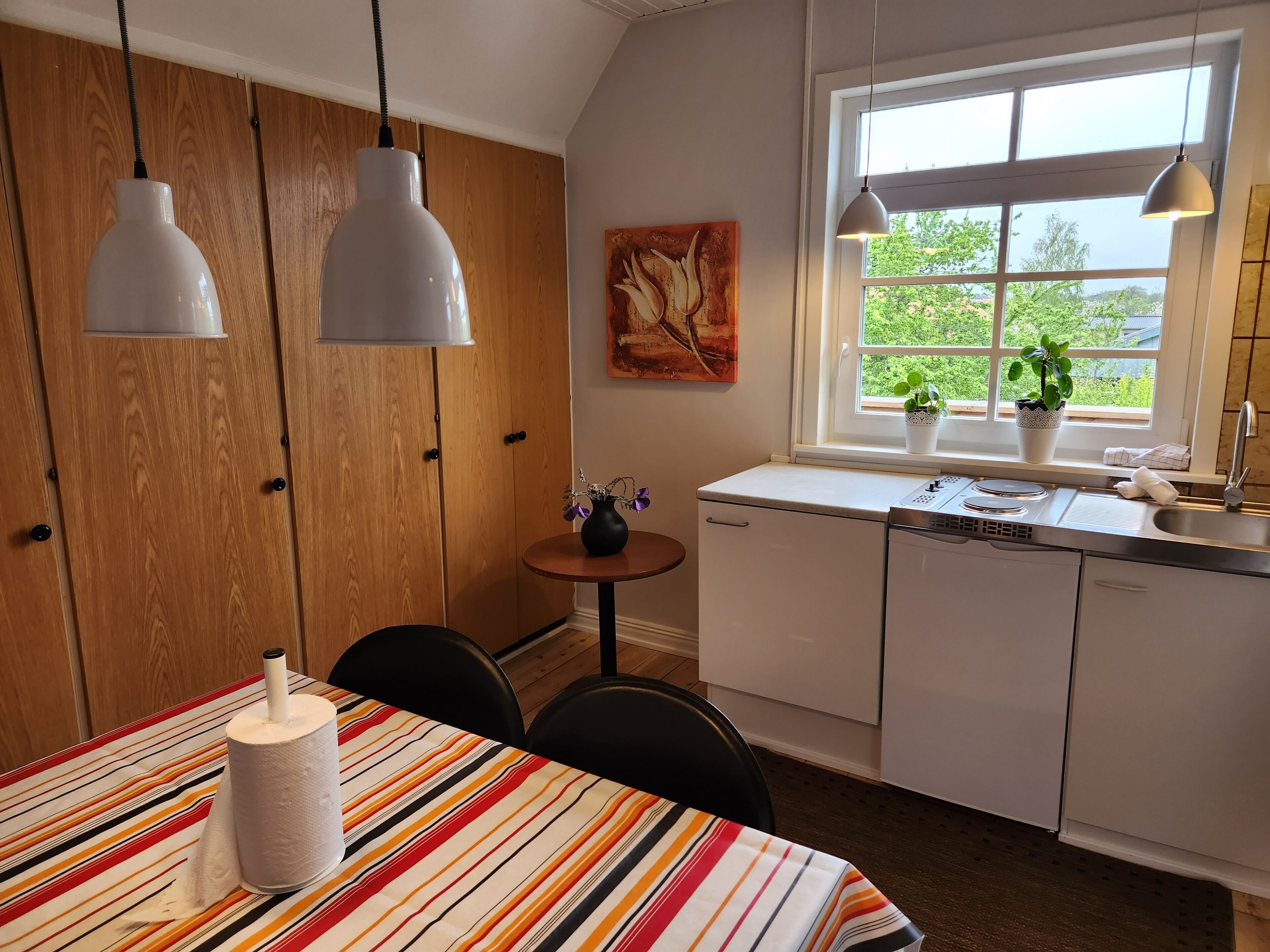 Une cuisine avec des meubles en bois, une table à manger recouverte d’une nappe à rayures, des suspensions lumineuses, une fenêtre ornée de plantes et un tableau accroché au mur.