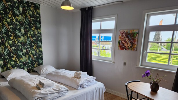 Een hotelkamer met twee bedden, een houten vloer, een tafel met stoelen, een raam met uitzicht op een weg en gebouwen, en een wand met een tropisch bladpatroon.