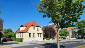 Een geel huis met een rood dak, een boom op de voorgrond en een rode auto geparkeerd op de straat.