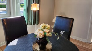Een eetruimte met een tafel gedekt voor twee personen, een vaas met bloemen, twee stoelen, een hangende lamp en een wandplank met foto's.