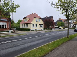 A two-story house with a red roof and a yellow facade, situated on a quiet street with a sidewalk and a grassy area.