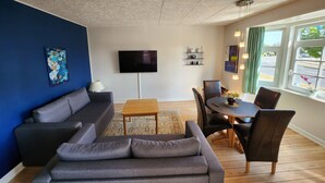 Een moderne woonkamer met een grijze bank, een houten salontafel en een eethoek met stoelen en een tafel. Een flatscreen televisie is aan de muur bevestigd, en er is een raam met gordijnen.