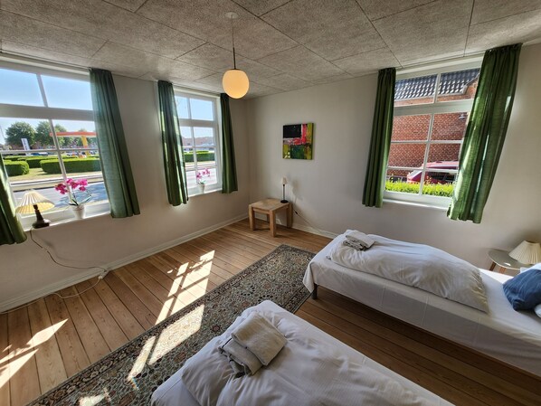 Et hotelværelse med to senge, et lille bord, en lampe, et vindue med gardiner og udsigt til det fri.
