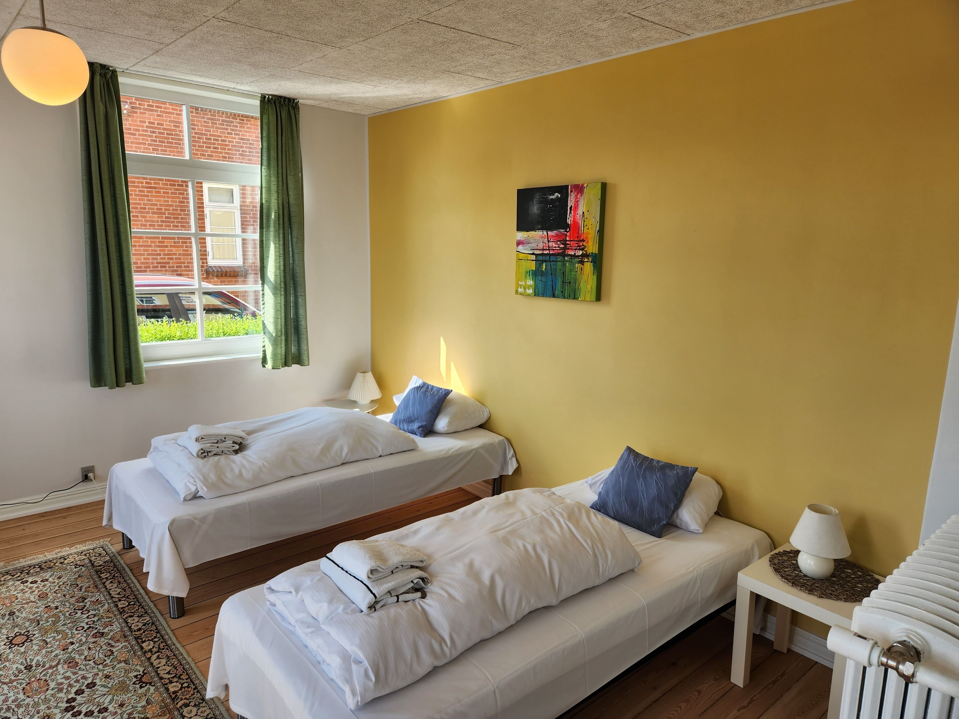 Habitación de hotel con dos camas, una ventana con cortinas, un radiador, una mesita con lámpara y un cuadro abstracto de colores en la pared.