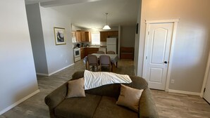 A living room with a brown sofa, a dining table, and chairs. There is a kitchen in the background with wooden cabinets and a refrigerator. - Home away from Home
Main Floor of Home (Three Hills)