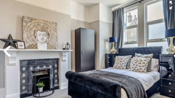 A bedroom with a fireplace, a bed with patterned pillows, a dark-colored headboard, and a nightstand with a lamp. - Margate Beach house (Cliftonville)