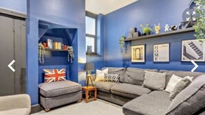 A living room with a grey sofa, a small ottoman, a bookshelf, and a Union Jack flag. - Margate Beach house (Cliftonville)