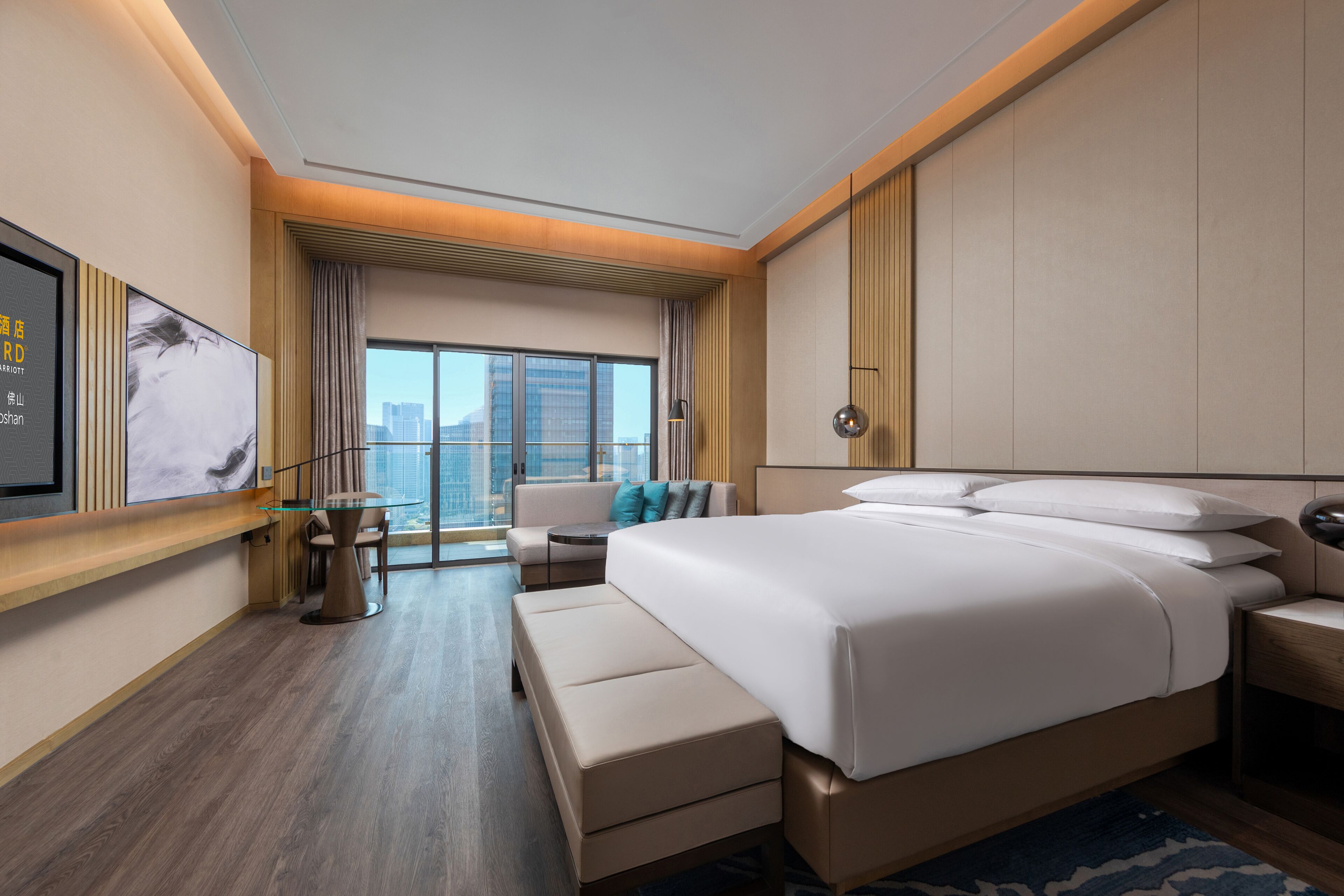 A modern hotel room with a large bed, a seating area with a sofa and armchairs, a small table, and a large window offering a city view.