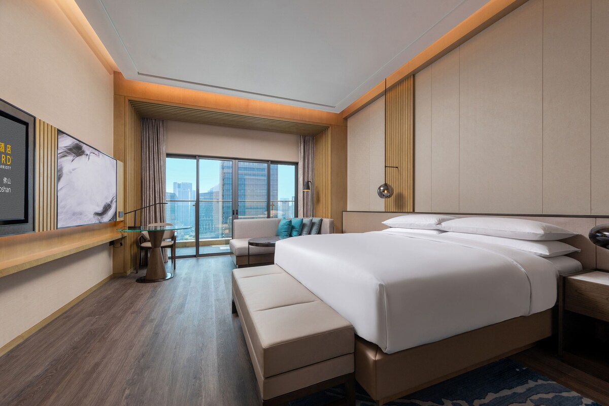 A modern hotel room with a large bed, a seating area with a sofa and armchairs, a small table, and a large window offering a city view.