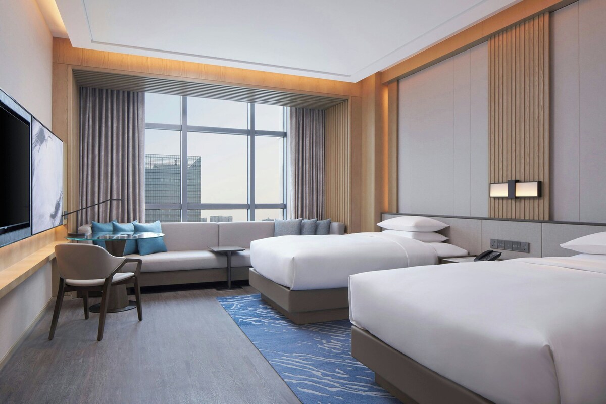 A modern hotel room with a large window, a sofa, two beds, a desk, and a flat-screen TV.
