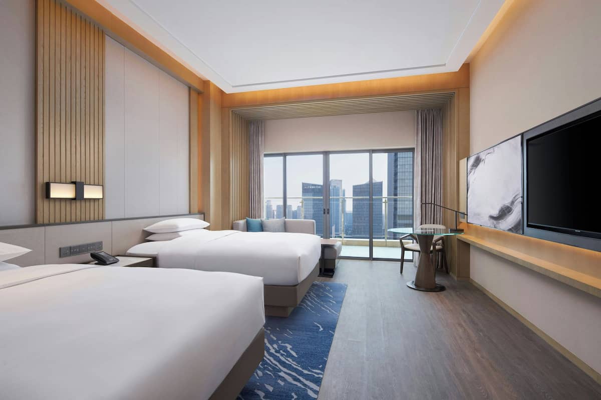 Habitación de hotel con dos camas, un televisor, un escritorio y un amplio ventanal con vista a la ciudad.