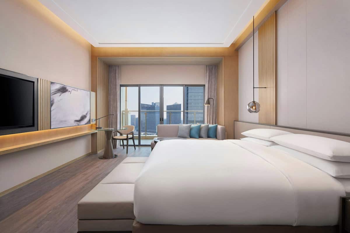 Una habitación de hotel moderna con una cama grande, un televisor de pantalla plana montado en la pared, una zona de estar con una silla y una mesita, y un ventanal con vistas a la ciudad.