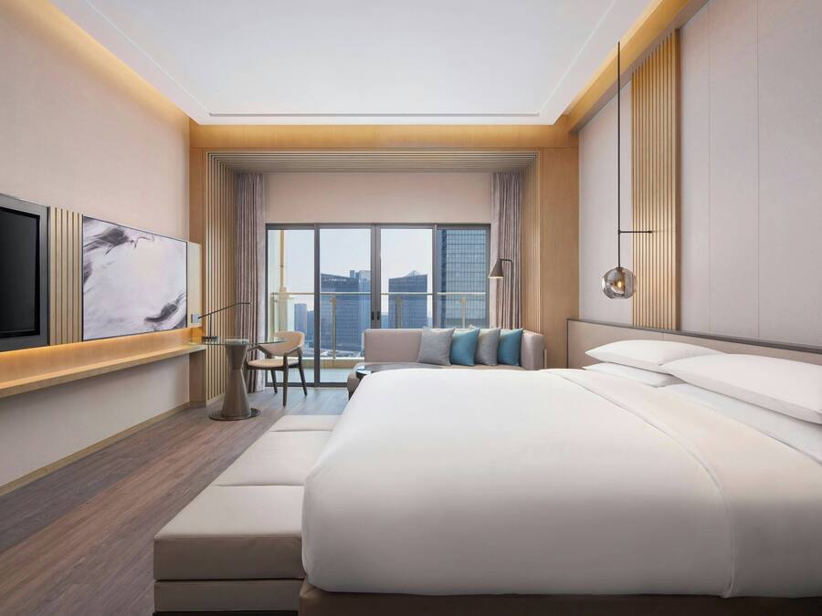 Una habitación de hotel moderna con una cama grande, un televisor de pantalla plana montado en la pared, una zona de estar con una silla y una mesita, y un ventanal con vistas a la ciudad.