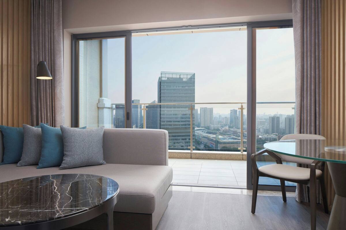 Una habitación de hotel moderna con un balcón grande, un sofá, una mesa de vidrio y una silla.