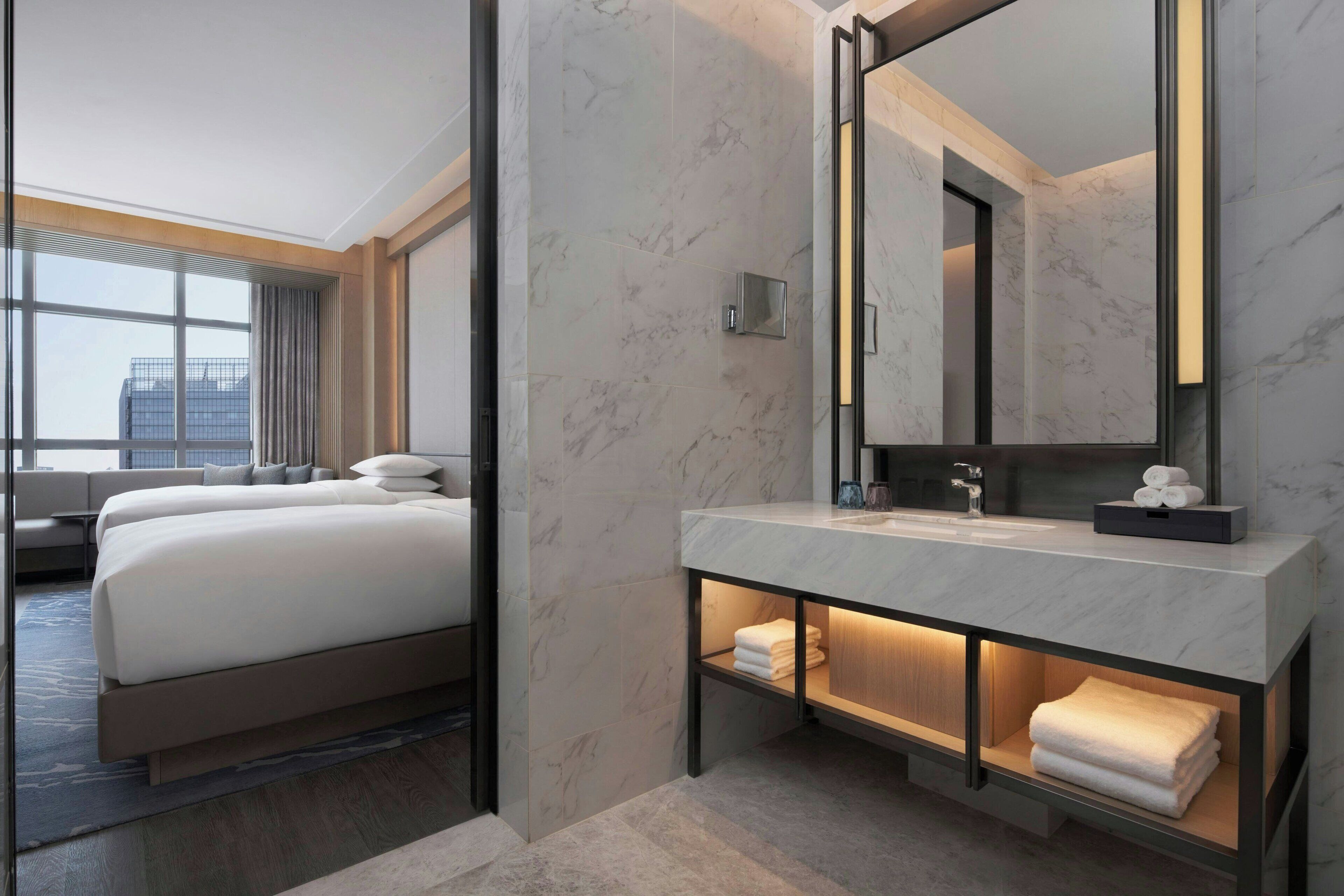 Una habitación de hotel moderna con una cama grande, un baño con encimera de mármol y un espejo amplio.