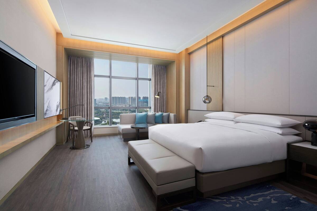 Una habitación de hotel moderna con una cama grande, un área de estar con una mesa y vistas a la ciudad desde la ventana.