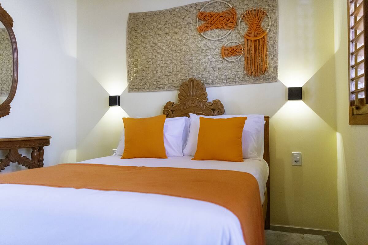 A neatly made bed with white and orange bedding, a decorative headboard, and wall-mounted sconces.