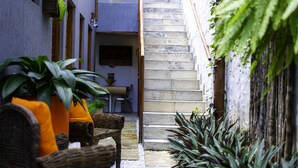 Um pátio estreito com uma escadaria que leva a um andar superior. Há móveis de vime e plantas em vasos.