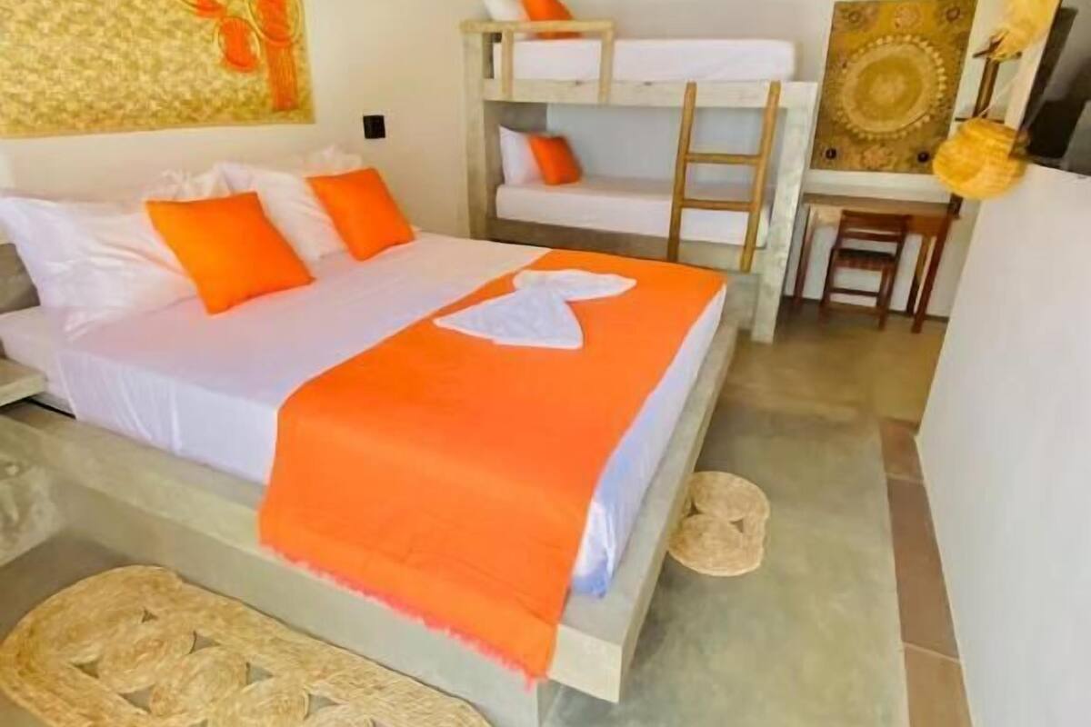 A bedroom with a bunk bed, a single bed with orange bedding, a woven mat, a wooden ladder, a wall art piece, and a hanging light fixture.