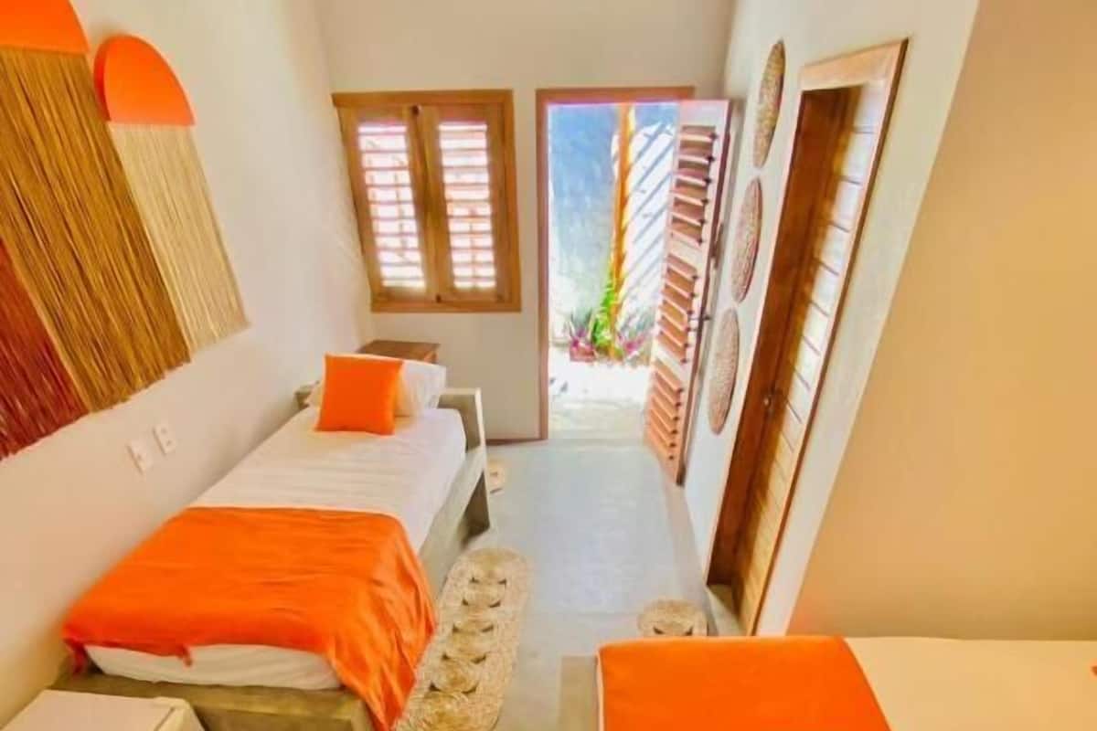 A hotel room with two beds, a small table, a suitcase, and a view of the outside.