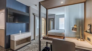 Una habitación de hotel moderna con un televisor de pantalla plana montado en la pared, un escritorio de madera con lámpara, una mesita de noche y un espejo grande que refleja la habitación.