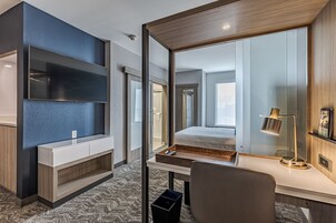 Una habitación de hotel moderna con un televisor de pantalla plana montado en la pared, un escritorio de madera con lámpara, una mesita de noche y un espejo grande que refleja la habitación.