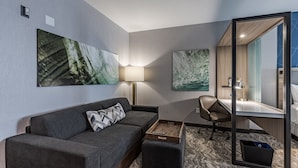 Moderni hotellihuone, jossa on harmaa sohva, työpöytä ja suuri peili. Seinillä on kaksi taideteosta, jotka esittävät luonnonmaisemia.