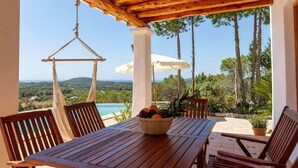 Una mesa de madera con un frutero, rodeada de sillas, bajo una pérgola de madera con vistas a una piscina y al paisaje.