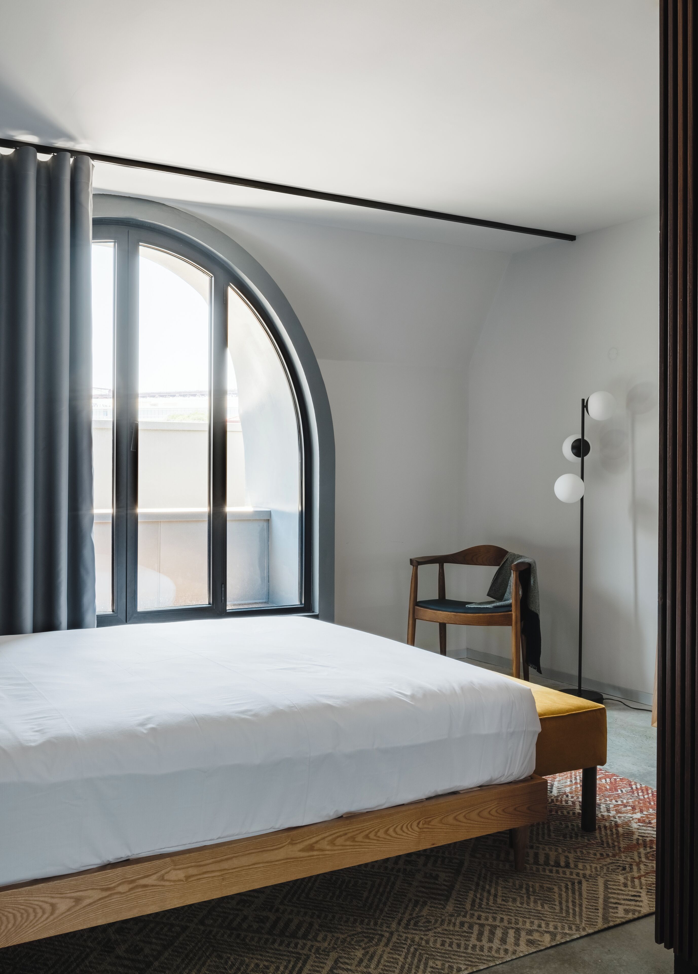 Ein Schlafzimmer mit einem großen Bett, einem Sessel, einer Stehlampe und Blick durch ein Fenster.