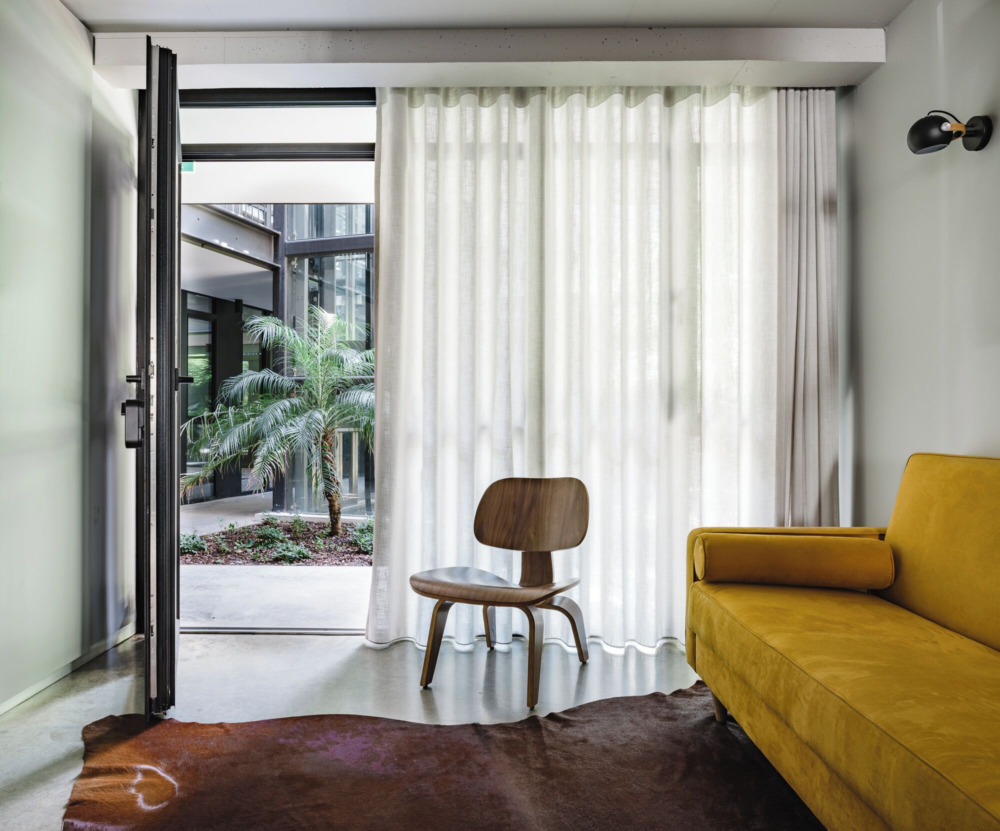 Uma sala de estar moderna com um sofá amarelo, uma cadeira de madeira e uma grande janela com cortinas leves.