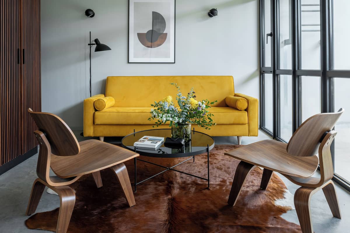 Un salon moderne avec un canapé jaune, deux fauteuils en bois, une table basse en verre et un tapis en fourrure.