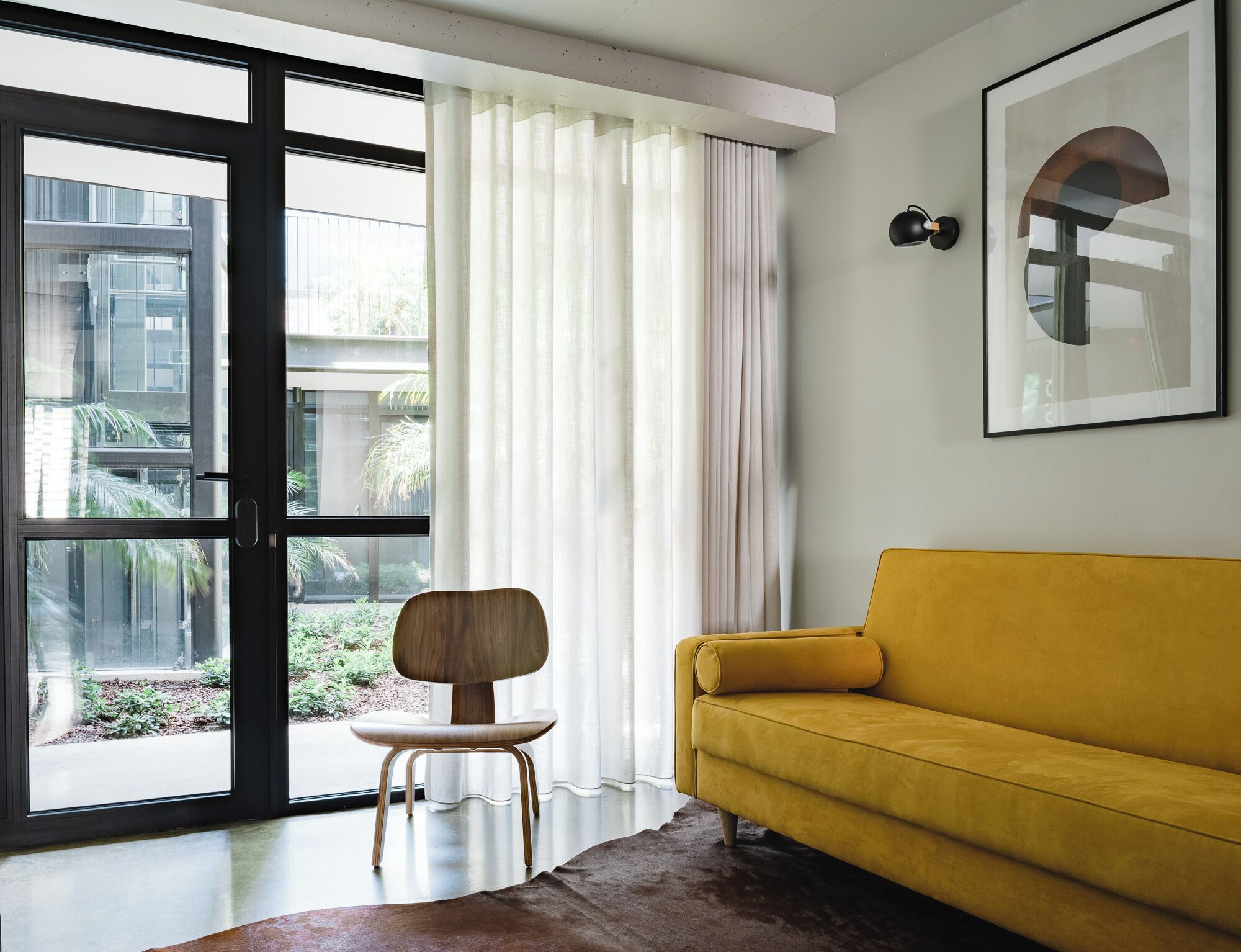 A modern living room with a yellow sofa, a wooden chair, and a large window with curtains.
