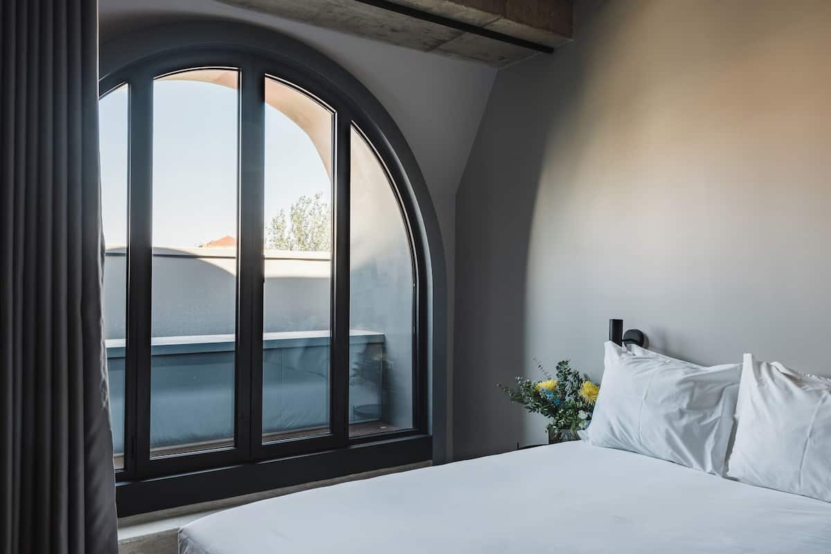 Une chambre à coucher avec une grande fenêtre en arc de cercle, un lit recouvert de draps blancs et un petit vase avec des fleurs.