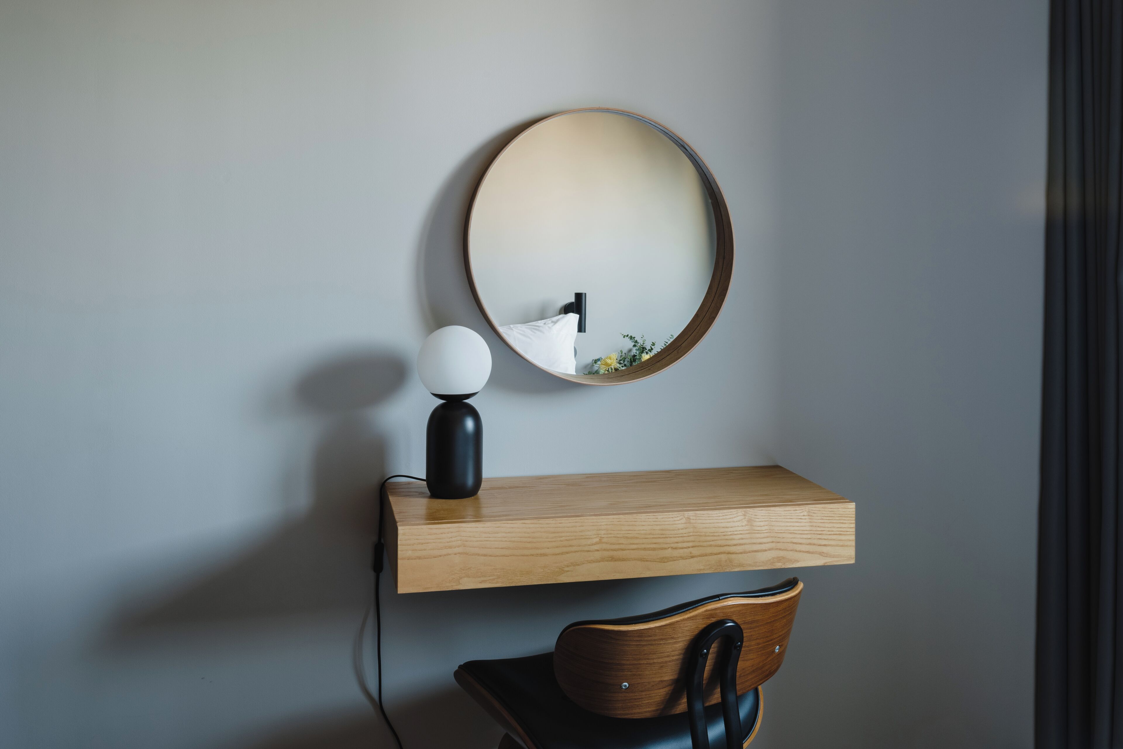 Uma mesa moderna com uma prateleira de madeira, um espelho redondo, uma luminária e uma cadeira.