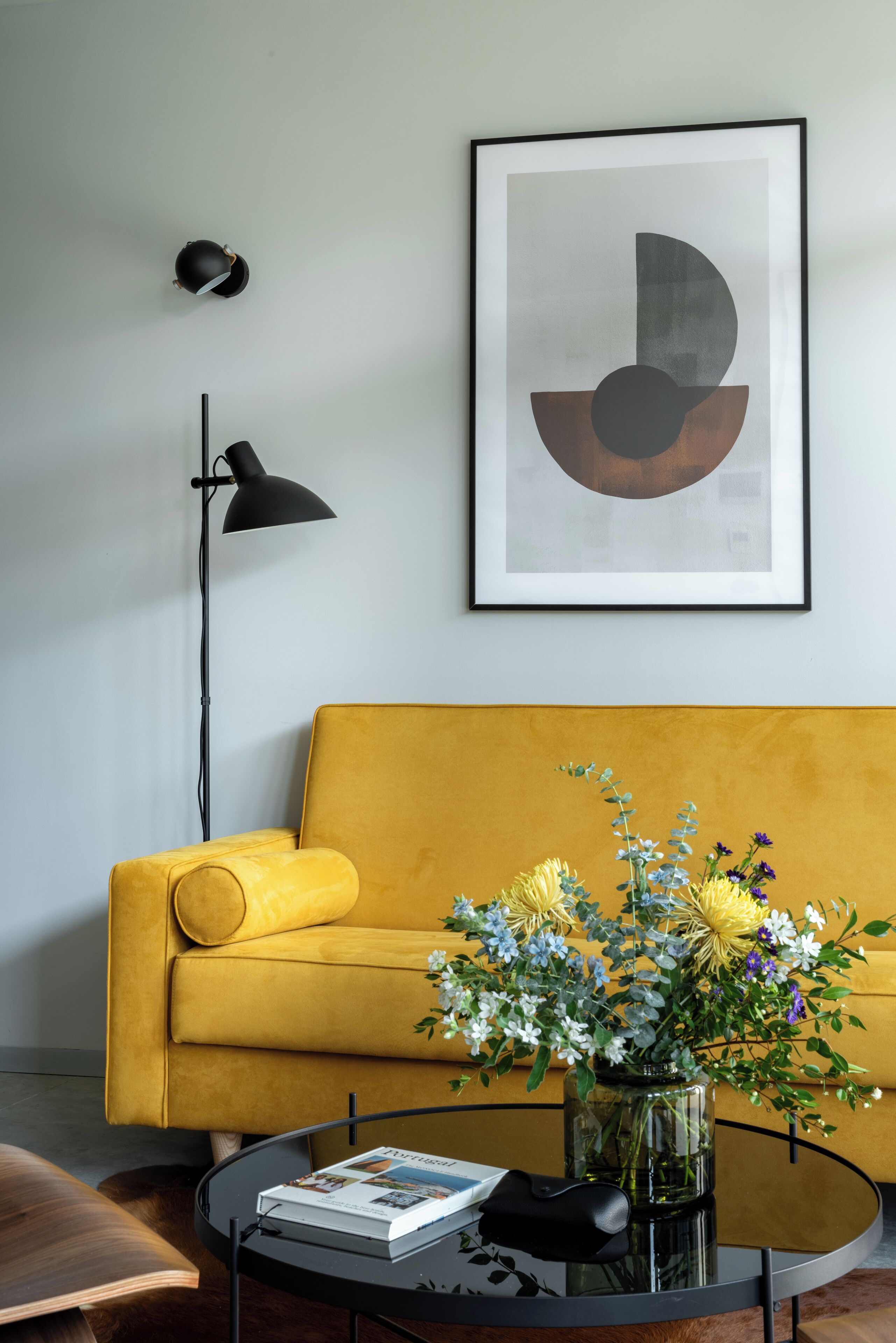 Een kamer met een gele bank, een zwart vloerlamp, een lijst met abstracte kunst, en een glazen tafel met een bloevas en een boek.