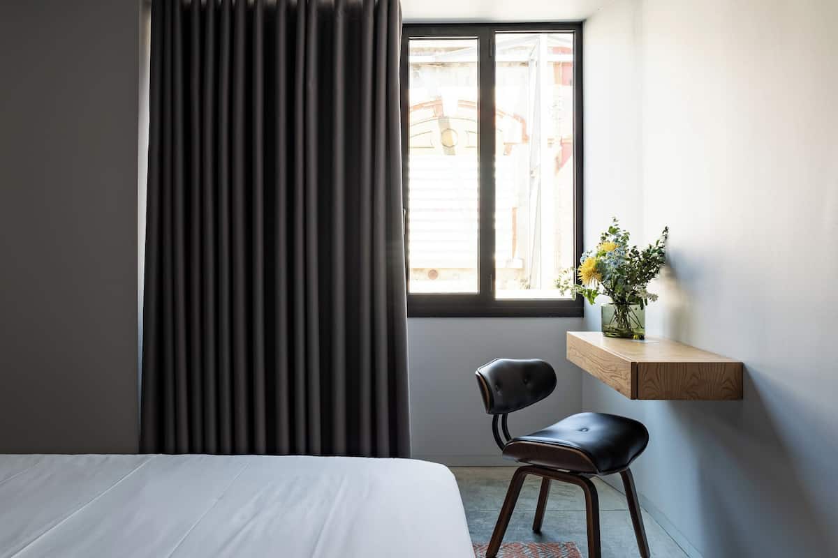 Une chambre d’hôtel avec un lit, une chaise, une fenêtre avec des rideaux et une petite étagère sur laquelle est posé un vase de fleurs.
