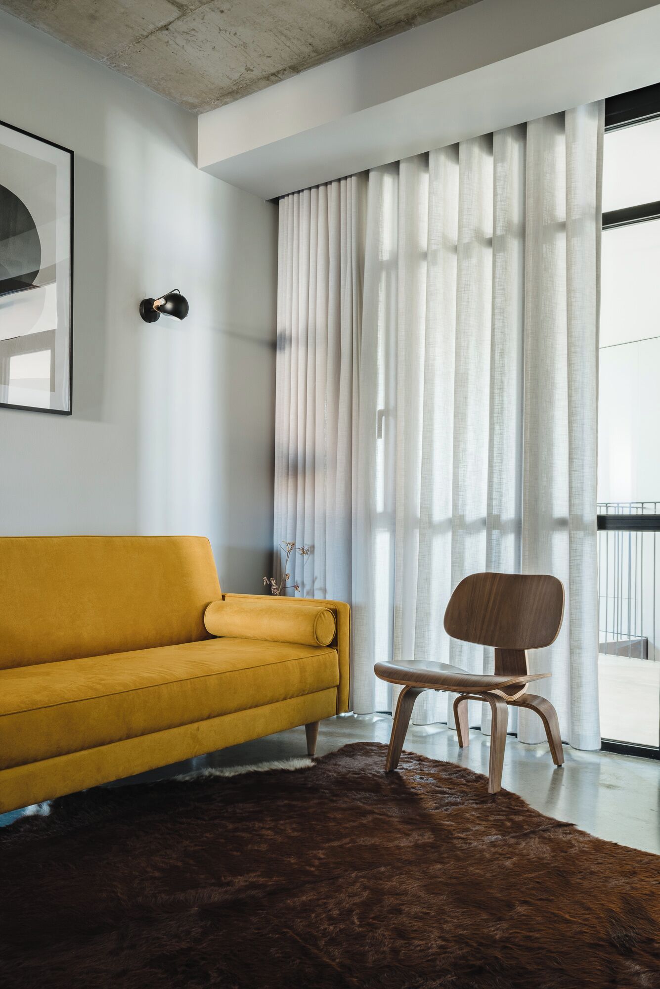 A modern living room with a mustard yellow sofa, a wooden chair, a large window with sheer curtains, and a framed picture on the wall.
