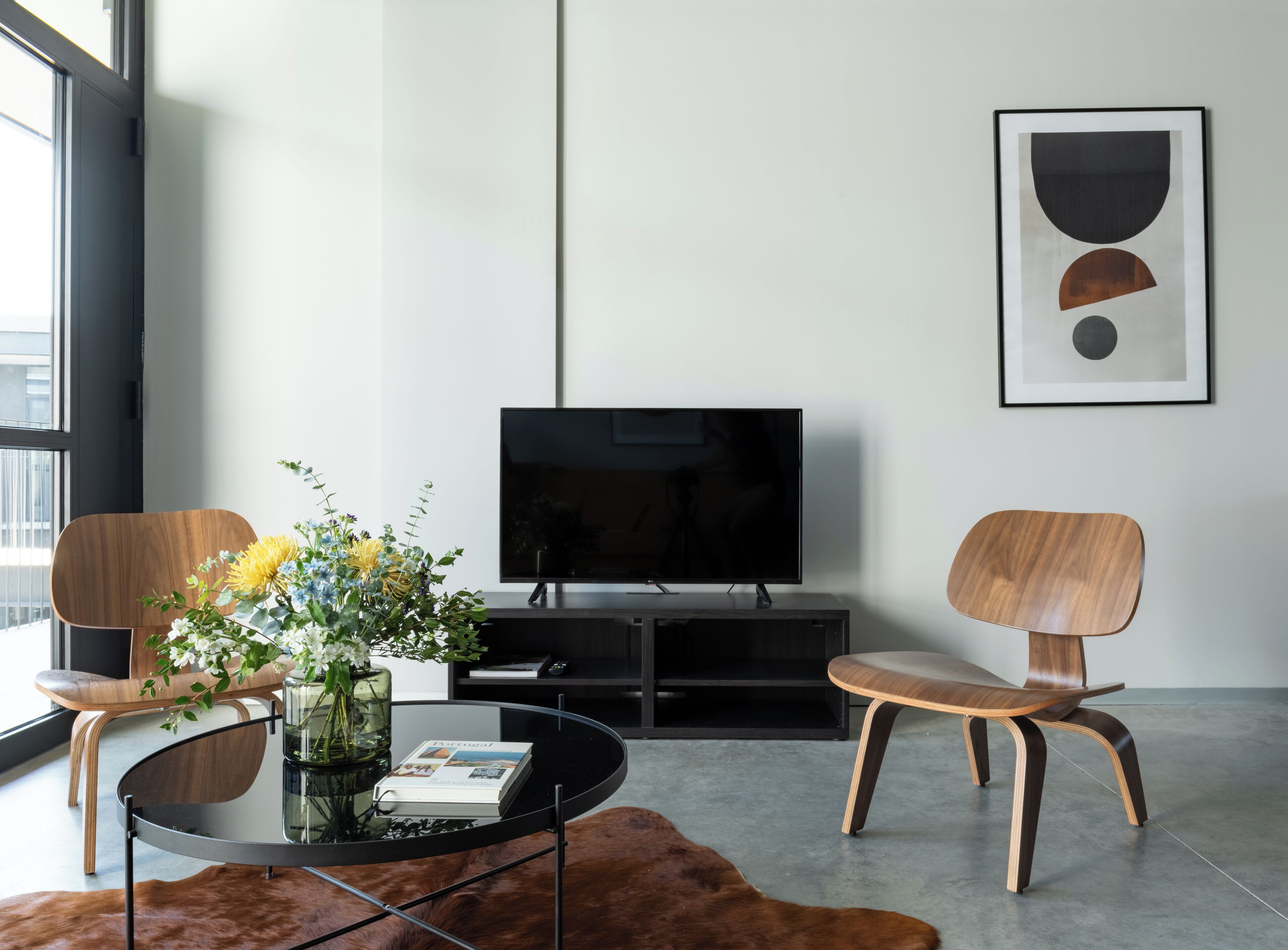 Un salon moderne avec un téléviseur à écran plat, deux fauteuils en bois, une table basse en verre, un magazine et une œuvre d’art abstraite encadrée accrochée au mur.