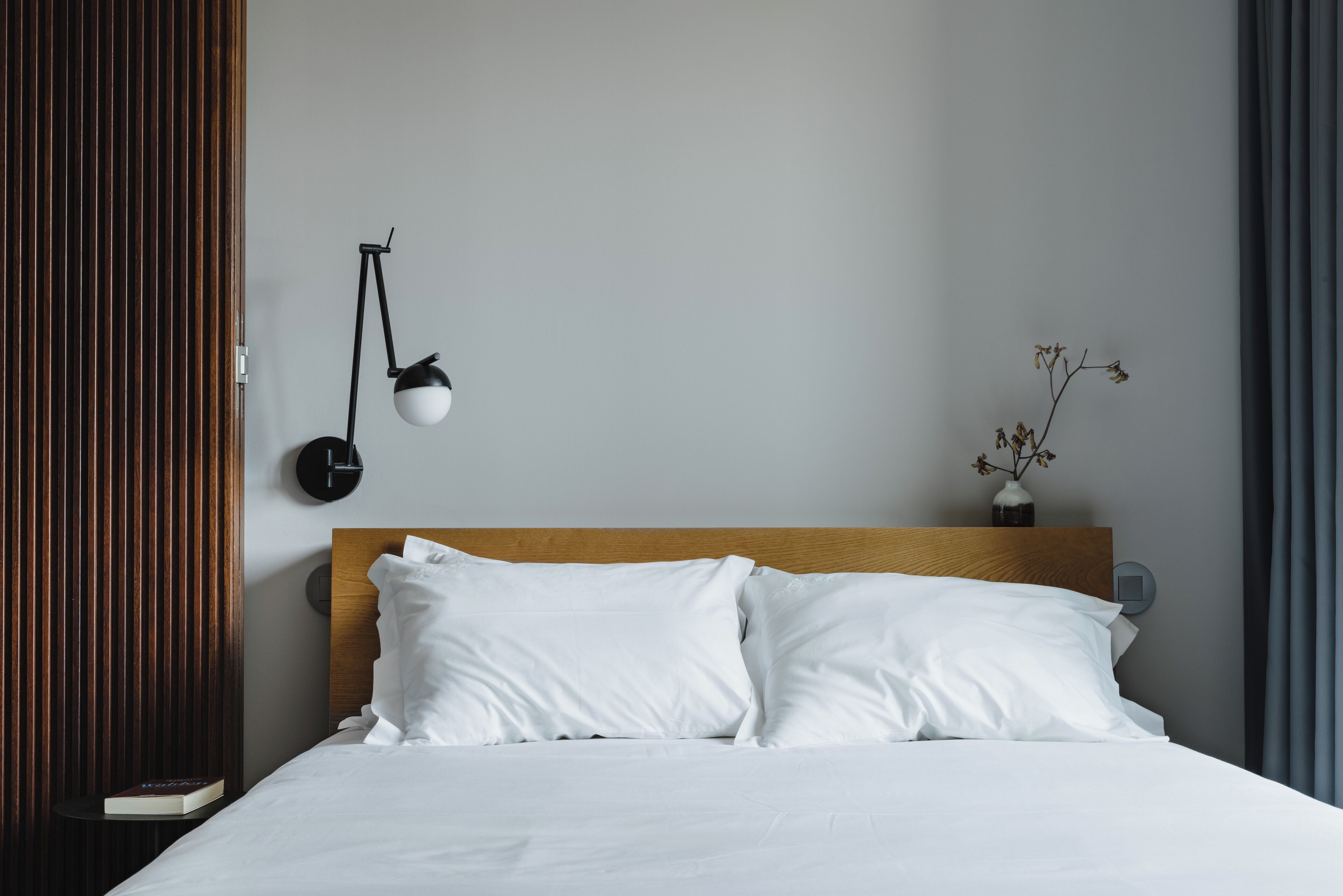 A neatly made bed with white linens, a wooden headboard, a bedside table with a book, a wall-mounted lamp, and a vase with dried flowers.